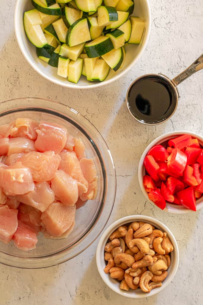 chicken breast in bite size pieces, green zucchini, soy sauce, red bell peppers, cashews in bowls 